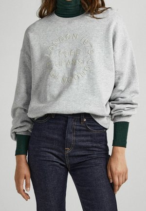 Personne portant un sweat-shirt gris clair avec le texte "PEPE JEANS LONDON 1973" en cercle, un jean bleu foncé et un col roulé côtelé vert en dessous.