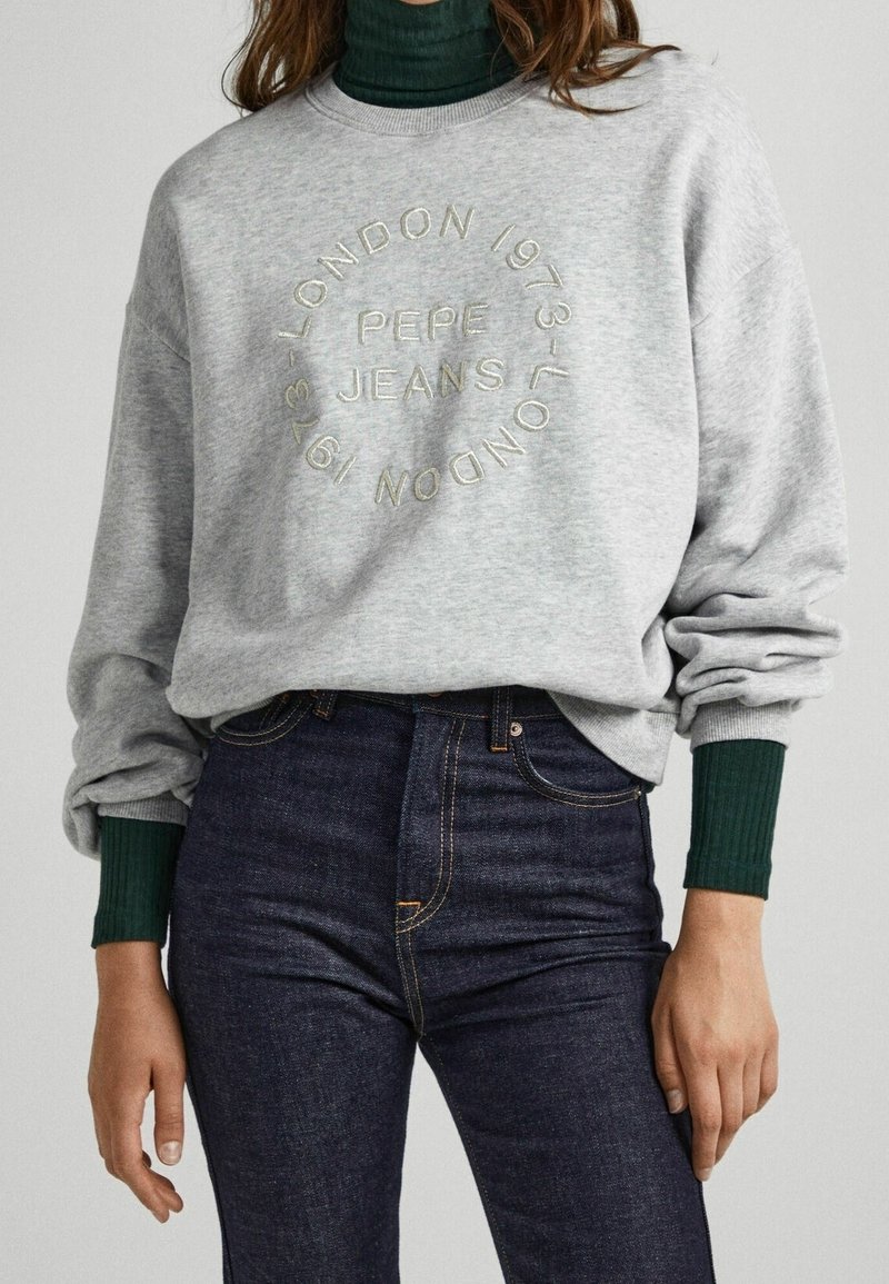 Personne portant un sweat-shirt gris clair avec le texte "PEPE JEANS LONDON 1973" en cercle, un jean bleu foncé et un col roulé côtelé vert en dessous.