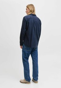 Man with shoulder-length blonde hair stands facing away wearing a dark long-sleeve shirt, blue jeans, and beige shoes against a plain background.