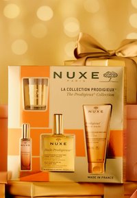 Coffret-cadeau NUXE comprenant une boîte dorée et orange, une bougie en verre transparent, une bouteille d'huile jaune et un tube de crème, tous ornés du logo NUXE.