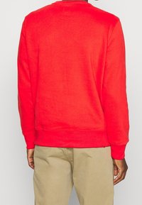 Uomo che indossa un semplice pullover rosso a maniche lunghe e pantaloni beigi, in piedi con la schiena rivolta verso uno sfondo neutro.