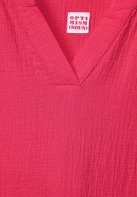 Tissu en coton rose avec une finition texturée, présentant un décolleté en V et une étiquette blanche avec "OPTIMISME (NOM)" imprimé en lettres rouges en gras.