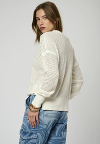 Pull en crème léger en tricot avec un motif en toile ouverte, des poignets et un ourlet côtelés. Présente une coupe décontractée et un design à épaule tombante.