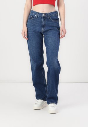 Femme portant un jean droit bleu, des baskets blanches et un top court rouge, debout devant un fond blanc uni.