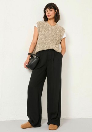 Beige gestricktes ärmelloses Oberteil über einem weißen T-Shirt, kombiniert mit schwarzen weiten Hosen und braunen Slippern. Schwarze Handtasche als Accessoire.