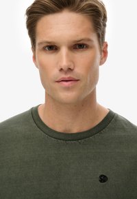 Oliven grøn bomuld t-shirt med rund halsudskæring og et lille sort logo på venstre bryst. Stoffet virker blødt med en glat tekstur.