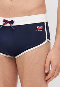 Tengerészeti úszóalsó fehér díszítéssel, piros és kék húzózsinórral, az "SPEEDO" logóval a bal oldalon. Simogató, rugalmas anyag.