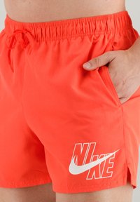Narancssárga fürdőnadrág, rugalmas derékkal, állítható zsinórral és fehér Nike logóval a bal alsó oldalon.