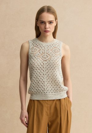 Femme aux cheveux blonds raides portant un haut en maille beige sans manches avec des motifs géométriques et un pantalon marron ample, debout devant un fond neutre.