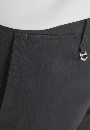 Close-up van de tailleband van zwarte, gestructureerde broek met een zilveren zeshoekige riemgesp en een wit, ingestopt topje.