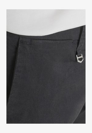 Close-up van de tailleband van zwarte, gestructureerde broek met een zilveren zeshoekige riemgesp en een wit, ingestopt topje.