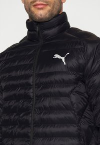 Sort pufferjakke med høj krave, quiltet tekstur, lynlås foran, og et hvidt Puma-logo på venstre bryst.