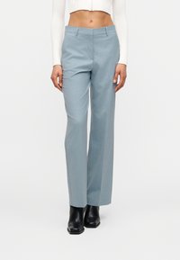 Γυναίκα που φοράει ανοιχτό μπλε παντελόνι καλοραμμένο, μαύρα μποτάκια μέχρι τον αστράγαλο και ένα κοντό λευκό μπλουζάκι με μακριά μανίκια, στέκεται μπροστά σε έναν απλό λευκό φόντο.