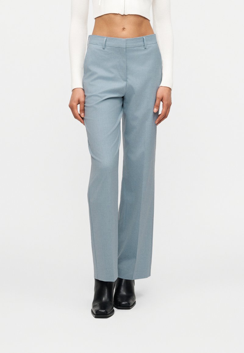 Γυναίκα που φοράει ανοιχτό μπλε παντελόνι καλοραμμένο, μαύρα μποτάκια μέχρι τον αστράγαλο και ένα κοντό λευκό μπλουζάκι με μακριά μανίκια, στέκεται μπροστά σε έναν απλό λευκό φόντο.