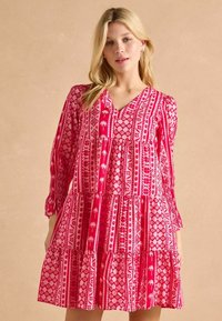 Różowa sukienka warstwowa z długimi rękawami, z dekoltem w kształcie litery V, zdobiona geometrycznym wzorem serc, palm i muszelek w różnych odcieniach różu.