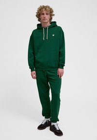 Zielona bawełniana bluza z kapturem, kangurzą kieszenią, ściągaczem na kapturem i małym logo. W zestawie z dopasowanymi zielonymi spodniami dresowymi i ciemnymi butami.