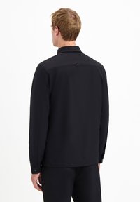 Man met kort lichtbruin haar, gekleed in een zwart langslavenshirt en zwarte broek, van achteren gezien tegen een witte achtergrond.