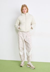 Giacca imbottita color crema con cappuccio, tessuto a trama, abbinata a joggers leggeri rosa con motivo e scarpe da ginnastica dorate, in piedi su pavimento verde.