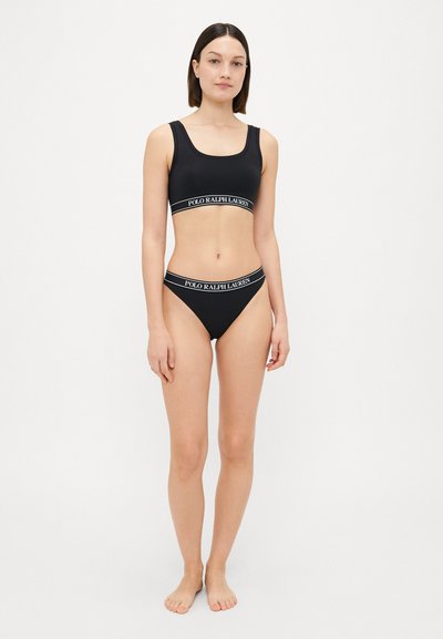 Melna sporta krūšturis un bikini komplekts no elastīga materiāla, iekļaujot baltu logotipa jostu. Gluda tekstūra, minimālisma dizains ar platām siksnām.