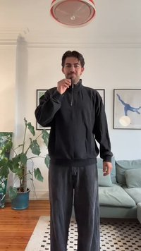 Felpa nera a zip con vestibilità rilassata, abbinata a pantaloni neri larghi; ambiente interno con piante d'appartamento e opere d'arte sullo sfondo.