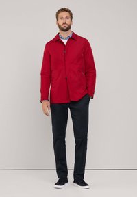 Giacca leggera rossa con chiusura a bottone e colletto a spread, abbinata a pantaloni blu navy e sneakers nere. Design semplice, texture liscia.