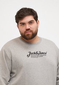 Sweatshirt gris avec un col rond et des manches longues. Présente le logo "Jack & Jones" en texte noir sur la poitrine. Texture de tissu douce.