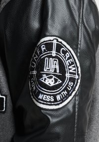 Svart skinnjakke med teksturert stofferm og et fremtredende rundt merke med hvit og grå broderi. Merket inkluderer teksten "UNFR CREW."