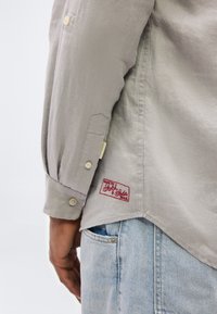 Κοντινό πλάνο του μπράτσου ενός ατόμου με μπεζ πουκάμισο με κουμπωτή μανσέτα και κόκκινη ετικέτα «Made by Scotch & Soda», δίπλα σε μπλε τζιν παντελόνι.