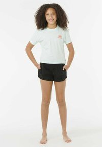 Chica con cabello rizado está descalza, vistiendo una camiseta azul claro con un pequeño gráfico de sol y pantalones cortos negros, con las manos en los bolsillos, sonriendo.