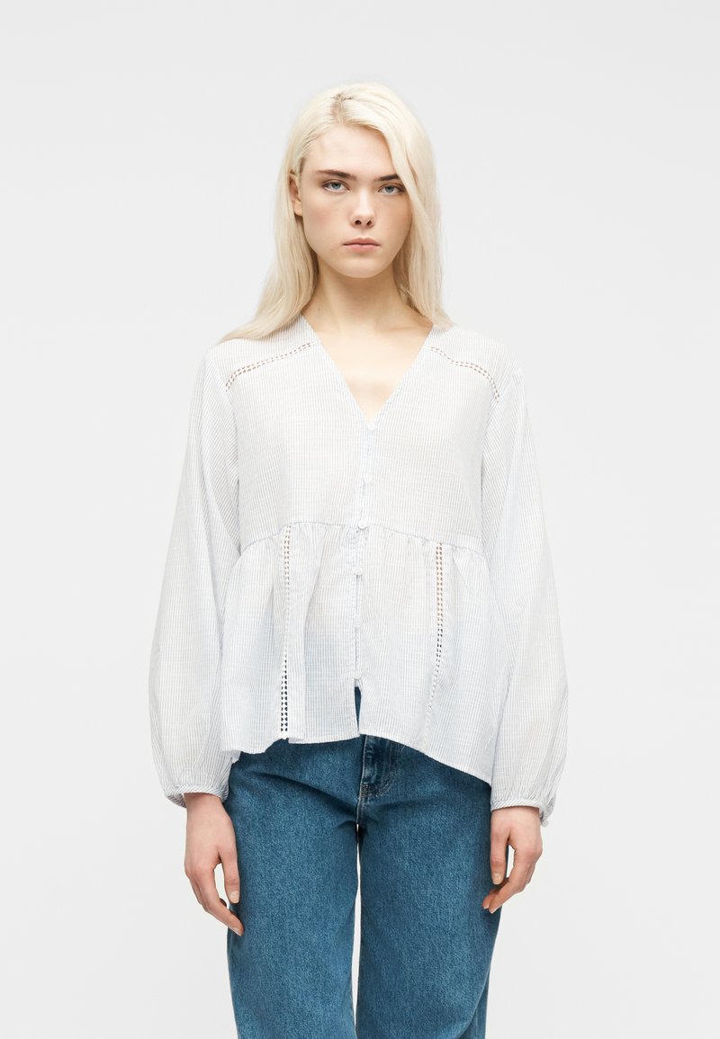 Νεαρή γυναίκα με μακριά ξανθά μαλλιά, φορώντας λευκό πουκάμισο με κουμπιά και μπαλούν μανίκια και μπλε τζιν, στέκεται μπροστά σε απλό φόντο.