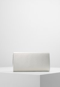 Bolso de mano rectangular plateado con una textura sutil y una cadena visible, colocado sobre un pedestal geométrico blanco contra un fondo liso.