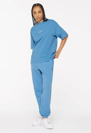 Blaues Baumwoll-T-Shirt mit kurzen Ärmeln und bedrucktem Text, kombiniert mit blauen Jogginghosen mit elastischen Bündchen und weißen Turnschuhen.