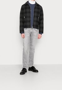 Černý a šedý károvaný kabát se zapínáním na zip, vrstvený přes námořnicky modré tričko, v kombinaci se světle šedými džíny a černými koženými botami.