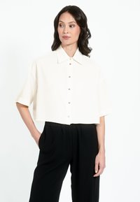 Cropped white shirt with short sleeves, featuring a button front and a classic collar. Paired with black, loose-fitting trousers.