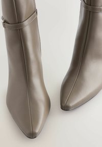 Bottines à talons hauts en cuir gris doux avec un bout pointu, présentant un détail de sangle sur le côté et une texture lisse.