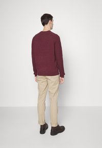 Bordeauxfarbener Strickpullover, beige Hosen, dunkelbraune Schuhe. Der Pullover hat gerippte Bündchen und Saum. Einfaches Design ohne sichtbare Muster oder Logos.