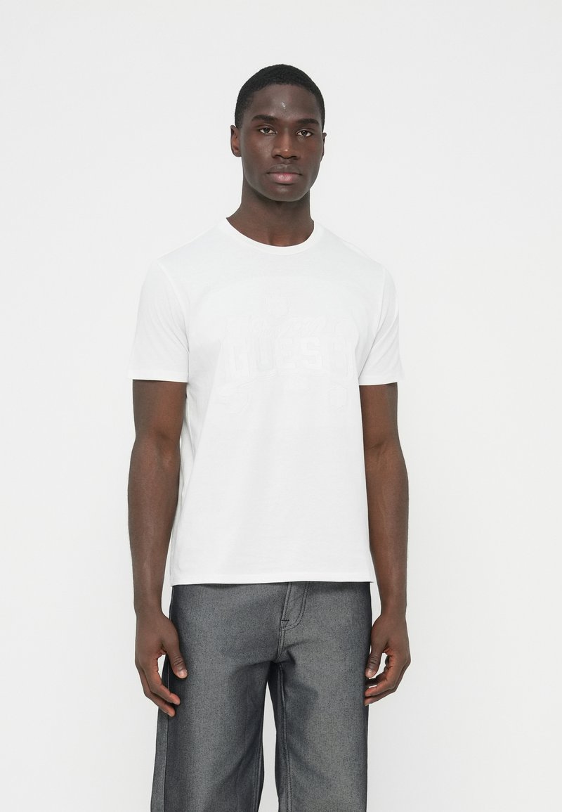 T-shirt bianca in cotone con maniche corte e scollo rotondo, con un sottile motivo grafico sul davanti. Indossata con pantaloni grigio scuro.