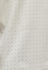 Šviesios spalvos audinys su tolygiai paskirstytomis mažomis juodomis dėmėmis ir centre atspausdintu tekstu bei logotipu „HKOUMORI“.