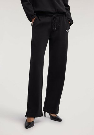 Persona con pantalones deportivos negros de pierna ancha con las manos en los bolsillos y zapatos de tacón alto negros puntiagudos sobre un fondo claro.