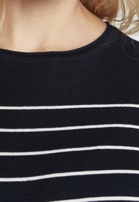 Námořnická modrá pletená halenka se bílými vodorovnými pruhy. Kulatý výstřih a žebrovaná textura. Jednoduchý design bez dalších vzorů nebo akcentů.