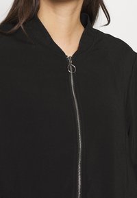 Giacca nera con zip, caratterizzata da una texture liscia, colletto a coste e hardware della zip in metallo. Il design sottolinea la semplicità e la versatilità.