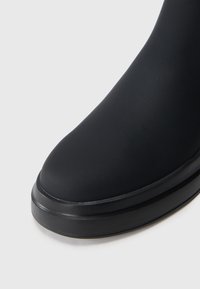 Sort ankelstøvle med en glat mat finish, rund tå og en tyk sort sål med diskrete vandrette riller for bedre fodfæste.
