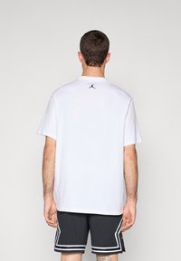 White cotton t-shirt with short sleeves, relaxed fit, features a small black Jordan logo on the upper back. Worn with black shorts.