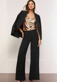 Sorte skræddersyet blazer over en guldpaliet V-udskåret top, parret med brede sorte bukser. Outfittet har en glat tekstur og elegant design.