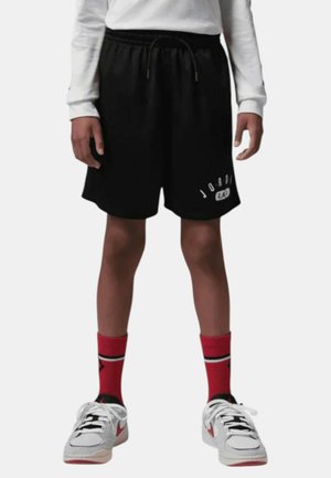 Personne portant un short Jordan 23 noir, un haut blanc à manches longues, des chaussettes rouges avec une bande noire, et des baskets blanches avec le swoosh Nike rouge.