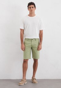 White short-sleeve t-shirt, light green shorts with pockets, and beige shoes with lace detail, standing against a neutral background.
