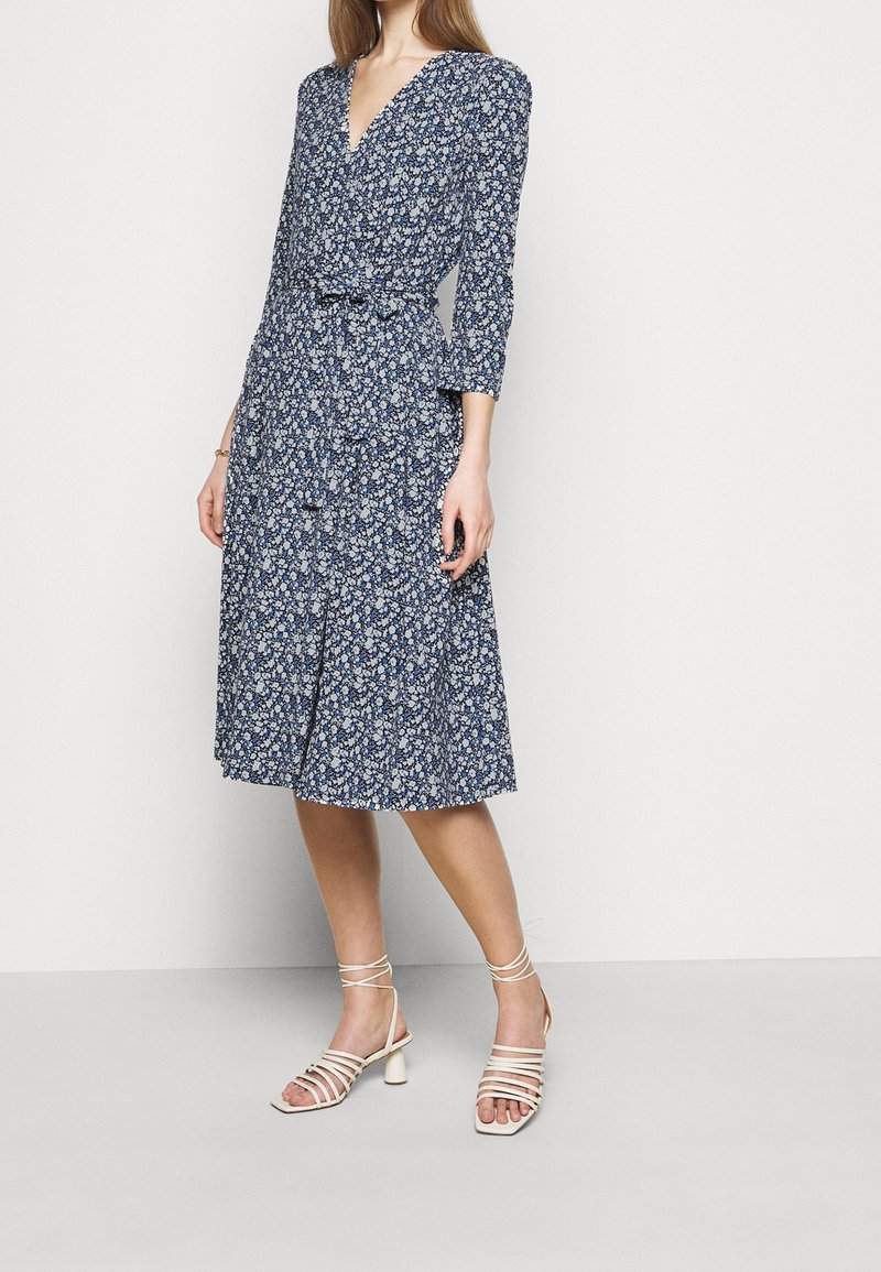 Femme portant une robe portefeuille bleue à motifs floraux, longueur genou, manches trois-quarts, et des sandales blanches à talons blocs avec des brides.