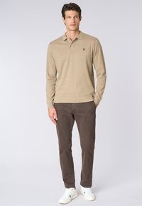 Uomo in piedi che indossa una polo beige a maniche lunghe, pantaloni marrone scuro e scarpe da tennis bianche, su uno sfondo chiaro e semplice.