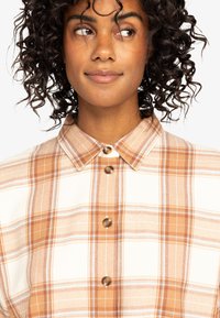 Button-up shirt with a plaid pattern in orange, beige, and white tones. Features a collar and brown buttons on a soft fabric.