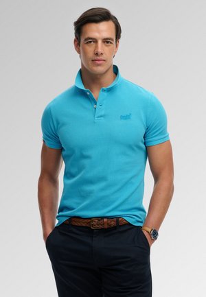Man die een felblauw poloshirt met korte mouwen draagt, donkere broek met een bruine gevlochten riem en een polshorloge, staande met zijn handen in de zakken.